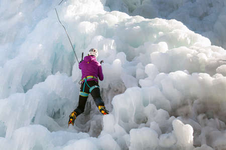 Female ice climber with ice climbing equipment, axes, helmet, harness, and crampons hanging on a frozen waterfall, back viewの写真素材