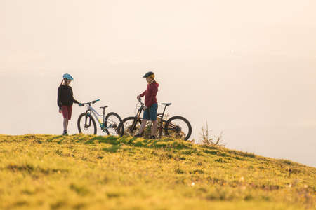 Two females on mountain bikes talking and looking at beautiful sunsetの写真素材