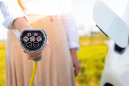 Close up shot of a females hand holding an electric car charger in the background visible car and...
