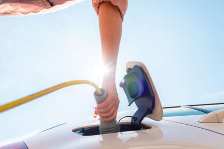 Low angle shot of a Caucasian woman opening an electric car charging socket cap and plugging in a...