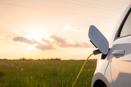 Electric car with opened charging socket cap and charger plugged in, at a public electric charging station near the highway at sunsetの写真素材