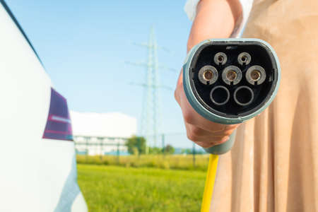 Close up shot of a females hand holding an electric car charger in the background visible car and electric power plantの写真素材