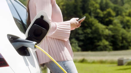 Woman browsing on a mobile phone while waiting to charge an electric car at a city public charger stationの写真素材