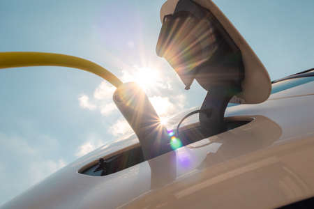 Woman plugging in electric car charger against sun and blue sky.の写真素材