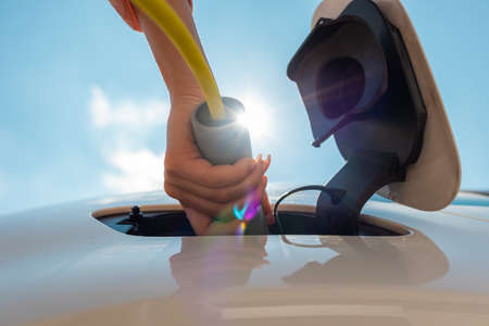 Woman plugging in electric car charger against sun and blue sky.の写真素材