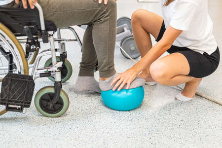 Pphysiotherapist and male patient seated in a wheelchair during rehabilitation treatment - press and release a foot on a small exercise ballの写真素材