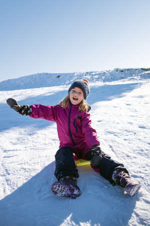 Llittle girl sledding with a warm knit hat, laughing descending the icy slope on the yellow shovel sledge.の写真素材