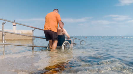 Man with disability on a wheelchair being transported into sea for swimming using a ramp.の写真素材