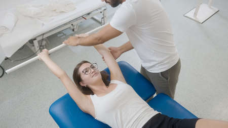 Woman doing exercises with medical bar, in a supine position. Male physiotherapist standing near her and assists in performing the exercisesの写真素材
