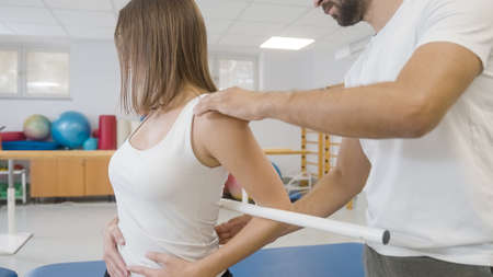 Woman during rehab treatment, and male physiotherapist helping her to do back and hip exercises with medical bar properlyの写真素材