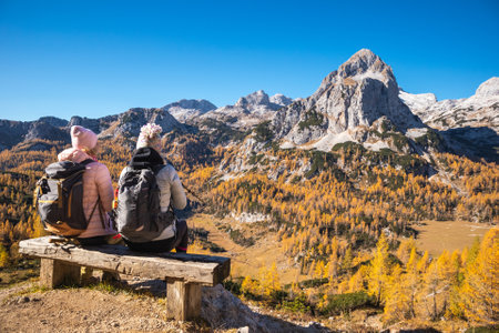 Two female hikers on a bench enjoying mountain panorama view on a sunny autumn day with golden larch trees.の写真素材