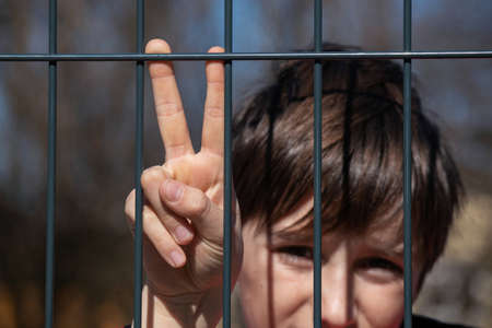 Child refugee showing peace sign behind a metal fence. Social problem of war migrants.