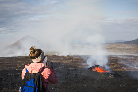 Woman tourist looking at dramatic view of dark volcanic landscape with glowing melting lava boiling in the crater, active volcano during eruption in Icelandの写真素材