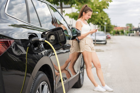 Young woman browsing on smartphone while waiting to charge an electric car in city. Renewable energy and new technology concept.の写真素材