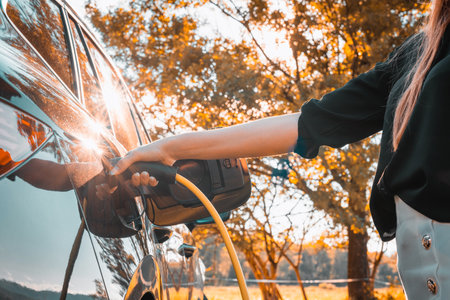 Female hand plugging in a charger in opened charging socket on a black electric car, with the sun shining through a green treetop in the backgroundの写真素材