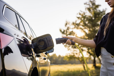 Female hand plugging in a charger in opened charging socket on a black electric car, with the sun...