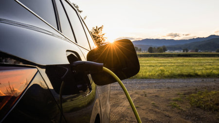 Black electric car with the power cable supply plugged in, in the blurred background visible green...