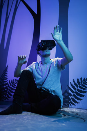 Young man wearing VR glasses sitting in a yoga pose on the virtual reality escape room floor. Fun,...