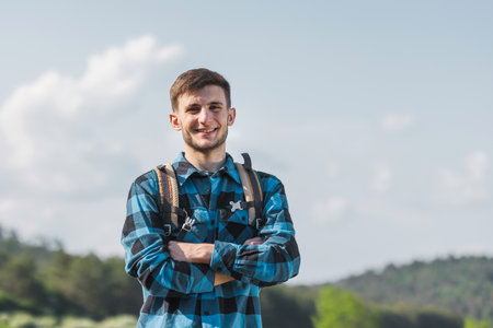 Portrait of a smiling, cheerful young hiker with a backpack on his back and a checked blue and black shirt, posing with beautiful nature in the background.の写真素材