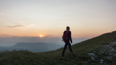 Female hiker silhouette with a backpack walking and following the trail uphill, enjoying the sky at sunset. Inspiration and healthy lifestyles concepts.の写真素材