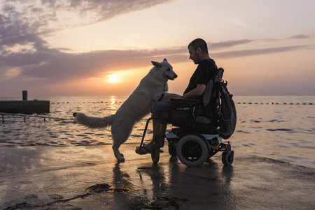 Man with disability, electric wheelchair user petting his dog and enjoying a sunset on the beach. People and animal friendship concept.の写真素材