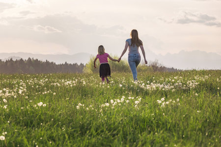 Mother and her little daughter running holding hands across the beautiful green grassland at sunset.の写真素材