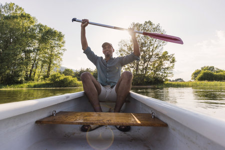 Cheerful young man sitting in a canoe on a calm lake and lifting a paddle above his head, front...