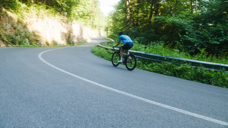 Male road racing cyclist in a blue sports jersey, with helmet and glasses, sprinting up a hill through the forest.の写真素材