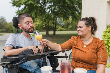 Caring wife helping her husband with a disability drink juice while enjoying sitting in the cafe garden. Love and commitment concepts.の写真素材
