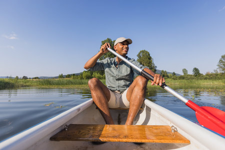 Young man laughing and having fun while canoeing on the calm lake surrounded by stunning nature, front view. Riverside leisure concept.の写真素材