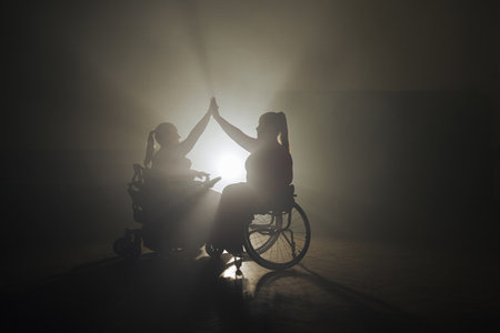 Two happy female para wheelchair dancers giving high five in a dance studio with dramatic backlit.の写真素材