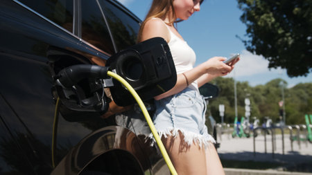 Beautiful girl plugging a cable charger into her parked black electric car and using a smartphone. Public electric vehicle charging station concept.の写真素材