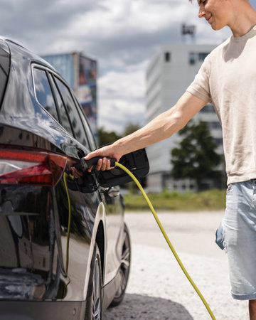 Man opening a socket and putting a charger in a black electric car at an EV charging station placed on city streetの写真素材