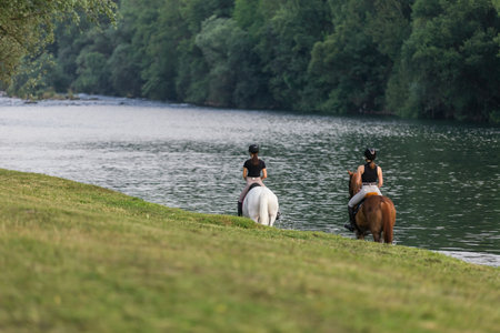 Riders, two young women riding beautiful horses down the calm river surrounded by the green groveの写真素材