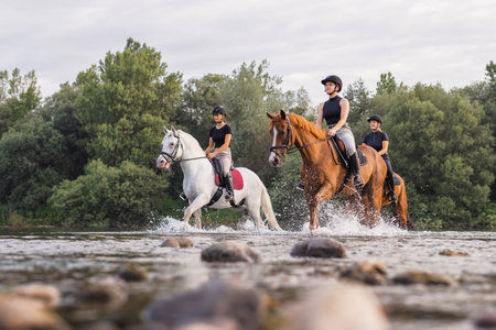 Three rider girls crossing the calm river water riding their beautiful horses on the cloudy summer day. Concept of equestrian leisure activity.の写真素材