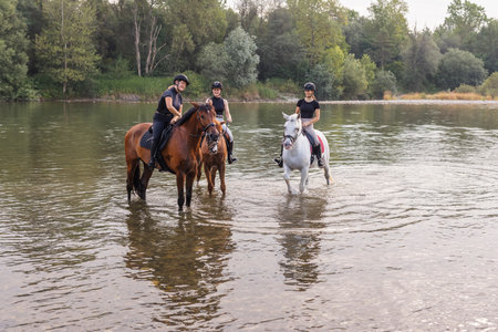 Three young women enjoying equestrian leisure, nature, and river, smiling while sitting on horsesの写真素材