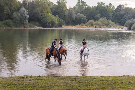 Three young women enjoying equestrian leisure, nature, and river, smiling while sitting on horsesの写真素材