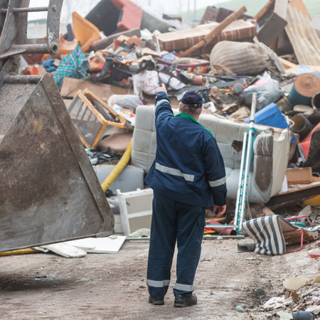Worker at a recycling center standing near a bucket loader, looking and pointing at the waste heap, rear view.の写真素材