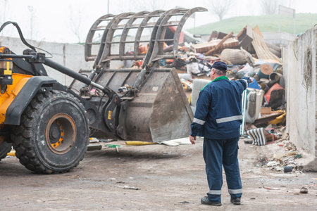 Male worker pointing on wheel loader and the garbage heap at a landfill site, rear view. Waste...