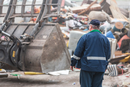 Man in a blue working suit looking at garbage near landfill skid steer loaderの写真素材