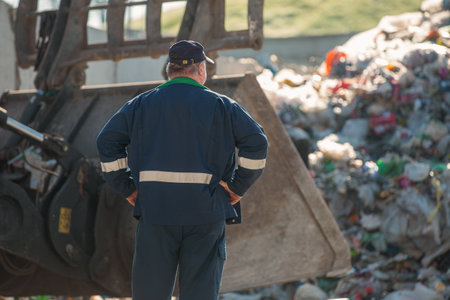 Man in a blue working suit looking at garbage near landfill skid steer loaderの写真素材