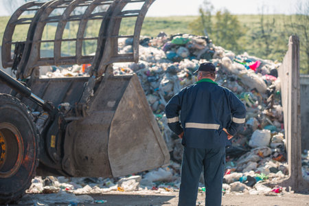 Recycling center worker, in dark blue work clothes, looking at an unsorted garbage heap just arrived...