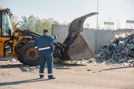 Landfill worker directing skid steer loader on the garbage heap, rear view. Waste disposal,...