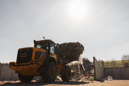 Yellow skid steer loader moving wooden waste material, shaking out a scrap grapple on the garbage heap in the materials recovery facilityの写真素材