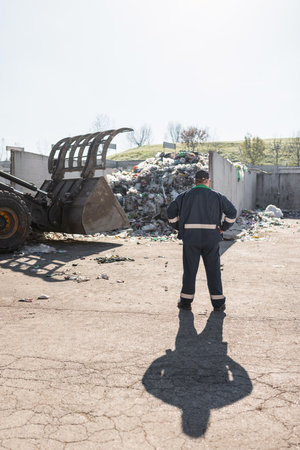 Recycling center worker, in dark blue work clothes, looking at an unsorted garbage heap just arrived...
