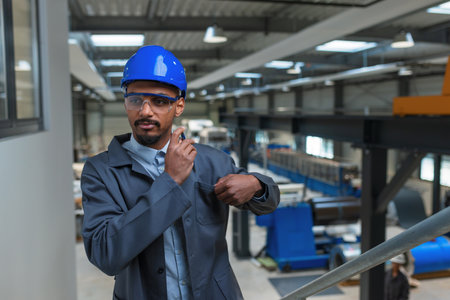 African American man with a blue helmet, a manager monitoring productivity in a factory and writing...