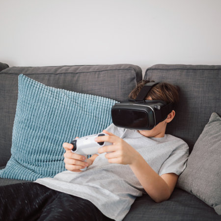 Caucasian male child wearing a VR headset and holding a gamepad while sitting in the living room,...