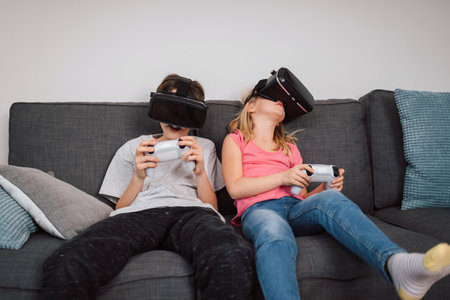 Two children, a boy and a little girl wearing VR headsets sitting in the living room playing in the...
