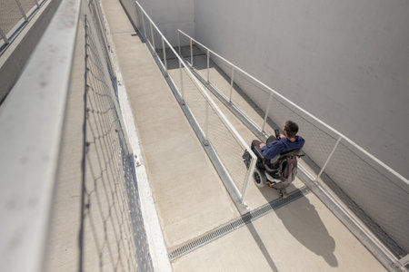 Man on electric wheelchair moving along concrete accessibility pathway near the building. Concept of wheelchair access facilitates.の写真素材
