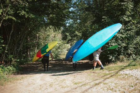 Three Caucasian recreational sportsmen going down to the river carrying kayaks and paddlesの写真素材